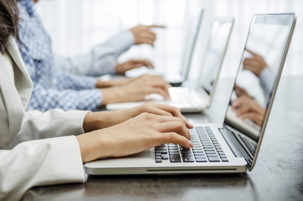 Laptops sitting on a table in a row with people typing on them. You can see 3 laptops and 3 sets of hands.