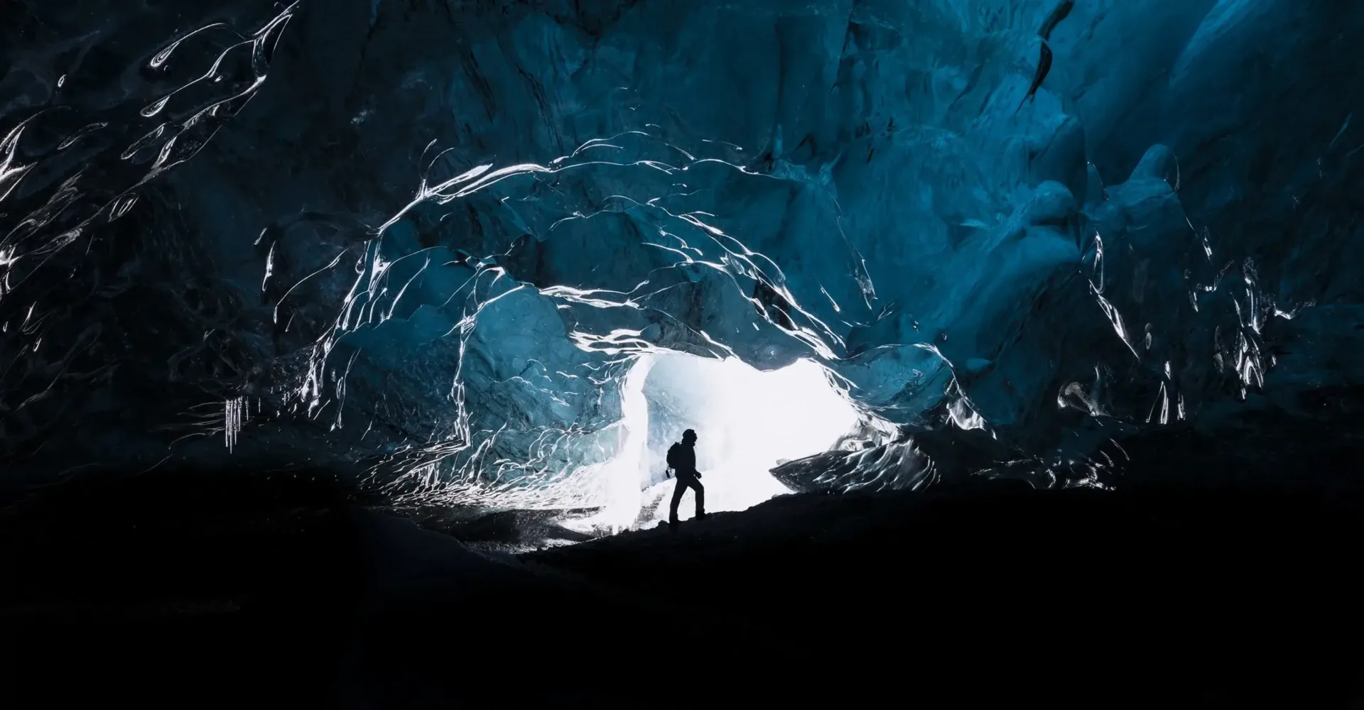 silhouette of a person standing at the entrance of a dark ice cave with glowing blue light behind them