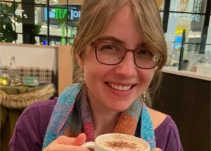 Smiling woman with glasses holding a cup of coffee in a cafe, colorful scarf around her neck.