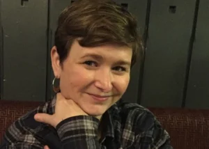 Smiling woman with short hair and hoop earrings rests her chin on her hands in a plaid shirt, seated in front of dark lockers.