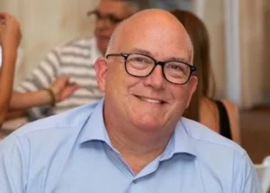 Smiling bald man with glasses in a light blue shirt at a social gathering.