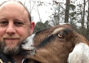 Bearded man taking a selfie as a brown-and-white goat nuzzles his cheek outdoors.