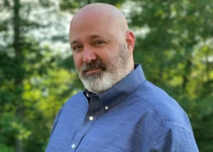 Portrait of a bald man with a gray beard in a blue button-down shirt outdoors, smiling slightly.