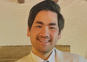Portrait of a smiling man with dark hair wearing a light beige suit and white shirt.