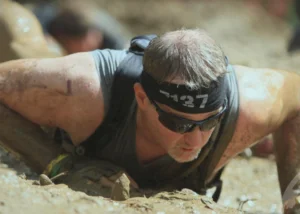 Male obstacle course racer crawling through mud, wearing headband with number 1437 and sunglasses on a sunny day.
