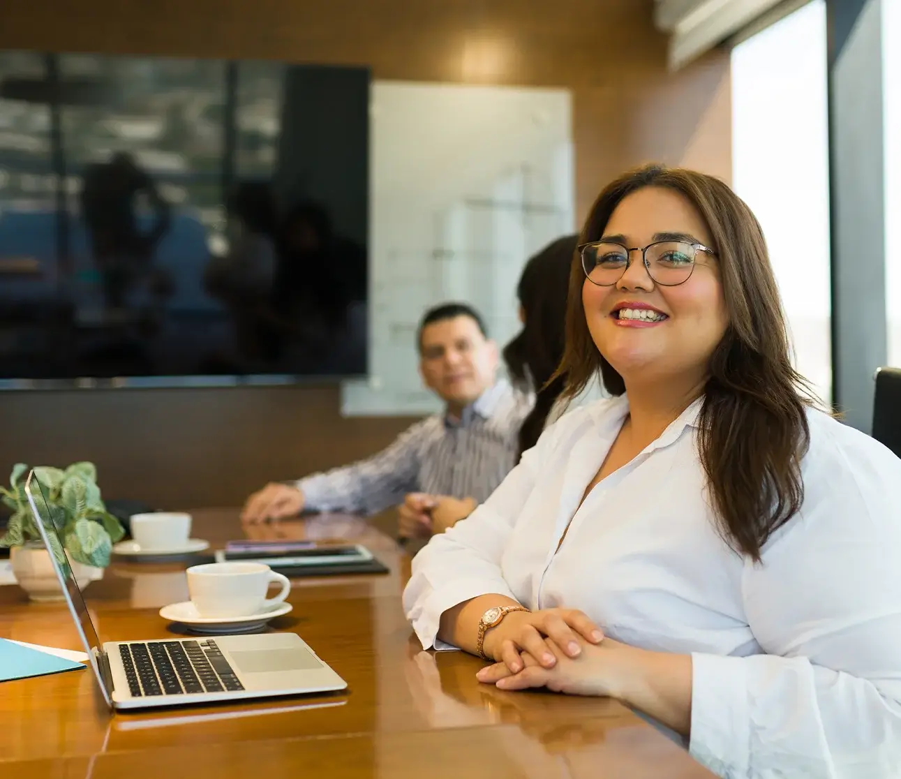 Smiling woman in a white blouse and glasses sits at a conference table with a laptop, coffee cup, and greenery; coworkers are in the background.