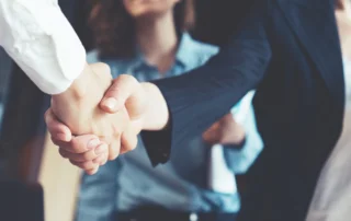 Two professionals shake hands in a blurred office setting, signaling an agreement or collaboration.