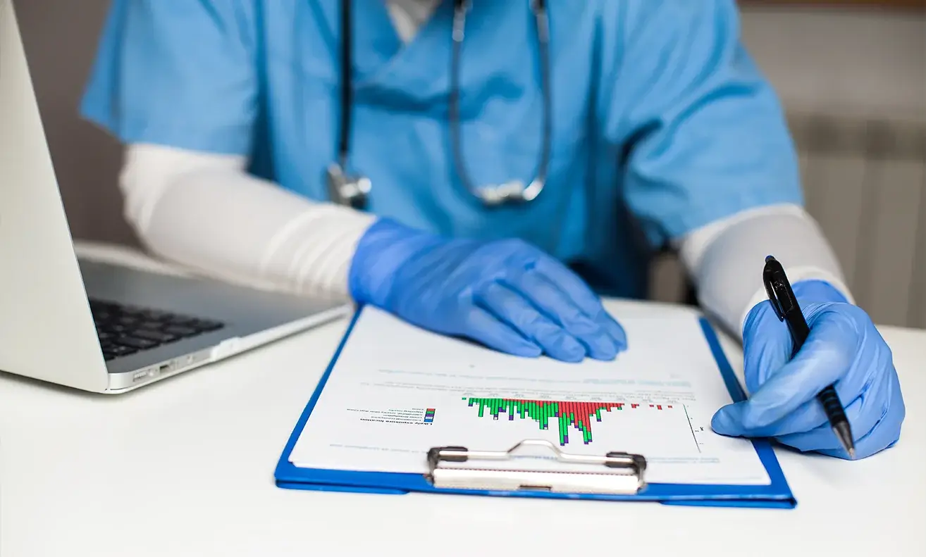 Medical professional in blue scrubs and gloves writes on a clipboard with a chart beside a laptop on a white desk.