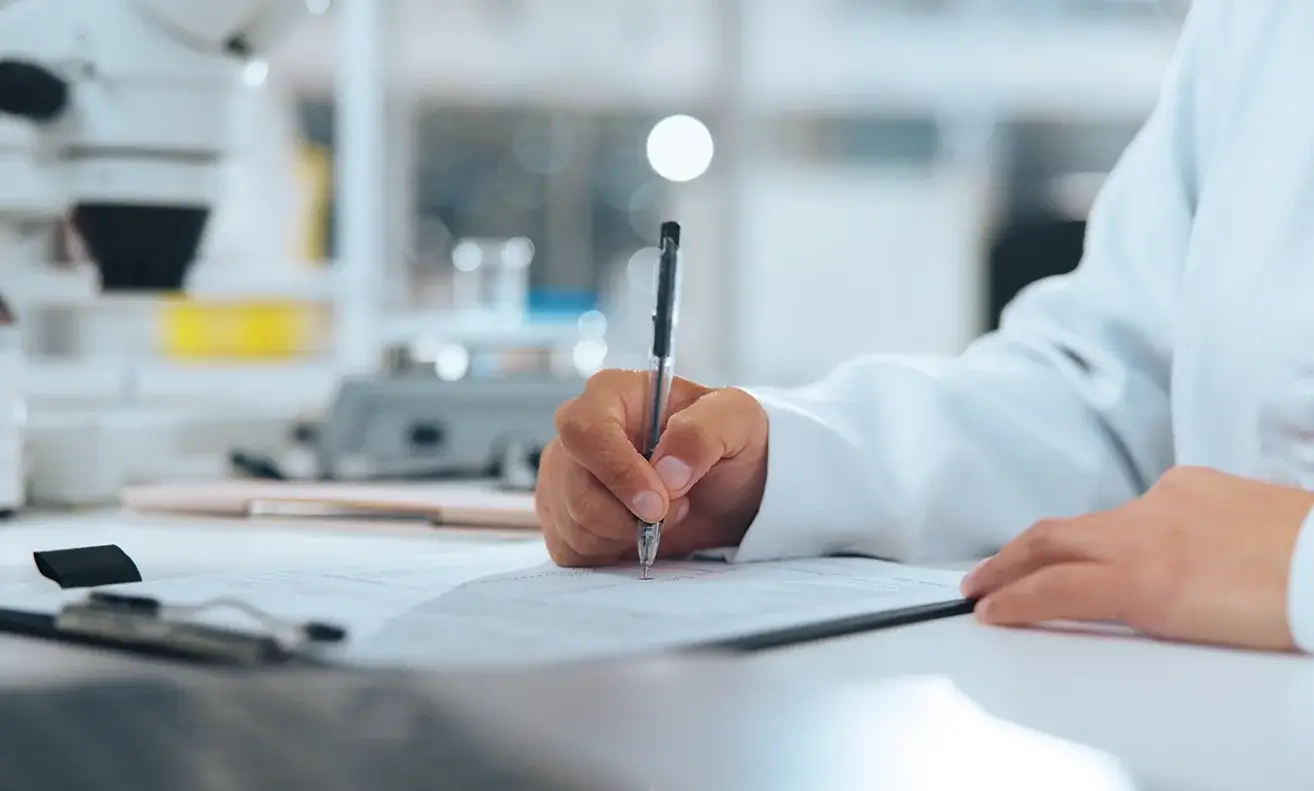 Scientist in a white lab coat writing on a clipboard in a laboratory setting.