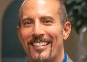 Portrait of a smiling man with short hair and a goatee, wearing a blue shirt and gray blazer.