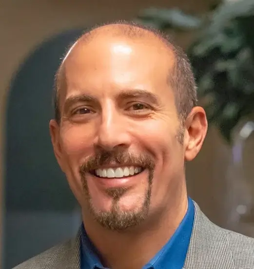 Portrait of a smiling man with short hair and a goatee, wearing a blue shirt and gray blazer.