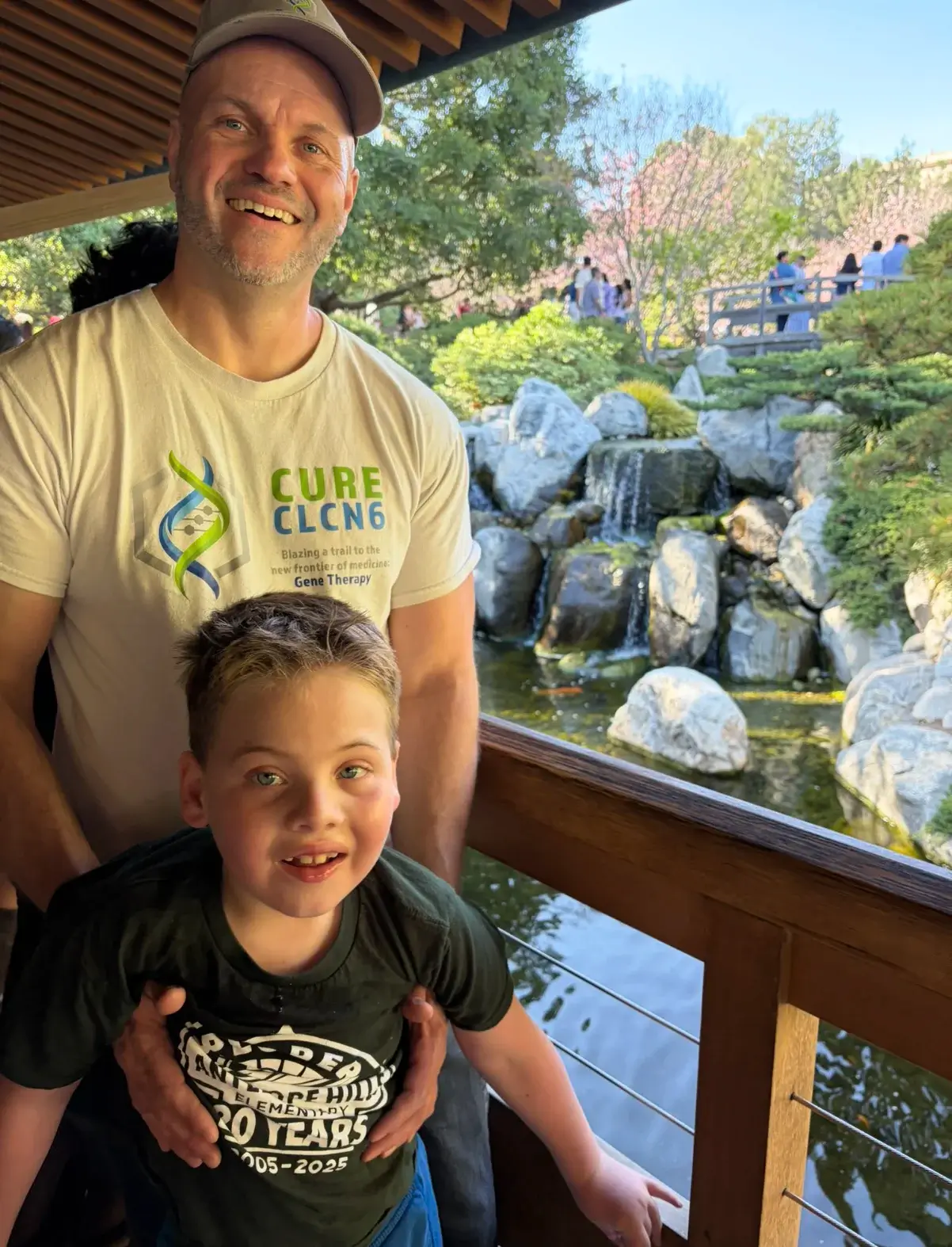 Smiling man in a beige CURE CLN6 T‑shirt and a boy stand on a wooden railing beside a rocky garden with a small waterfall in the background.