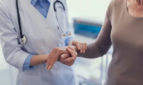 Doctor in a white coat with a stethoscope gently supports a patient’s hand during a consultation.