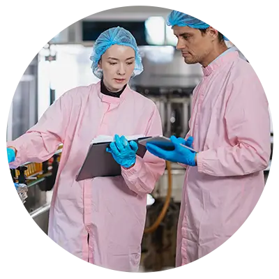 Two workers in pink protective gowns and blue gloves review data on a tablet in an industrial cleanroom setting.