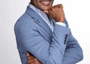 Smiling man in a light blue blazer with glasses, resting his chin on his hand in a professional pose.