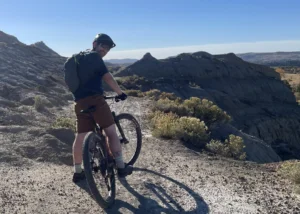 Mountain biker with helmet and backpack on a rocky trail overlooking a canyon under a clear blue sky, sunlit rocks in the distance.