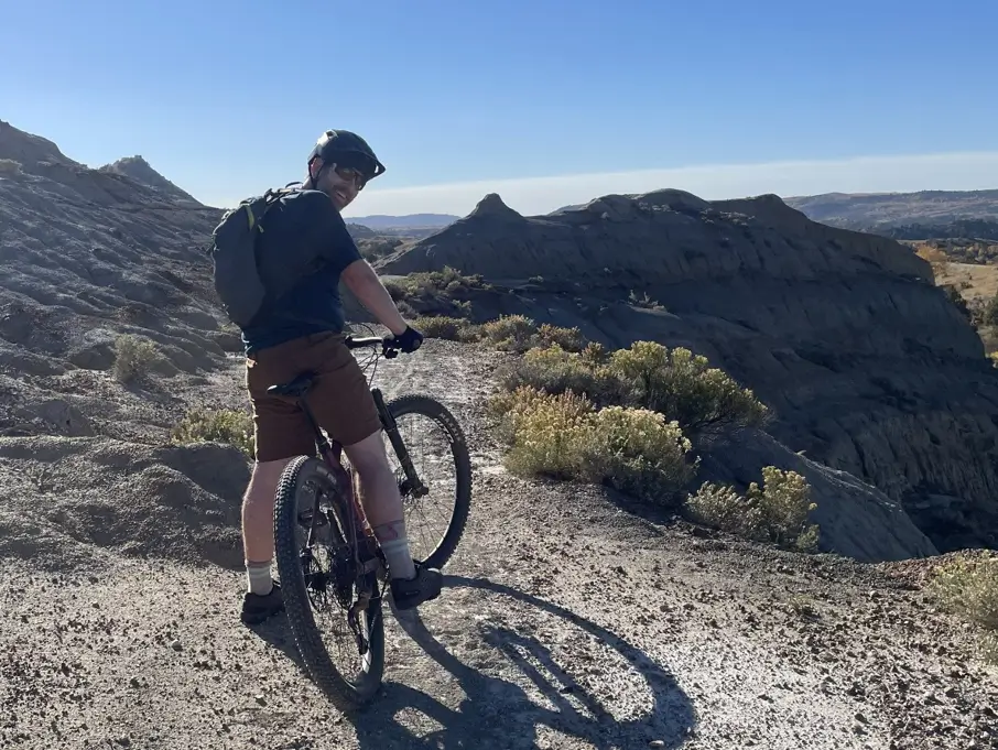 Mountain biker with helmet and backpack on a rocky trail overlooking a canyon under a clear blue sky, sunlit rocks in the distance.