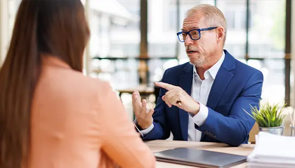 A man in a blue suit and glasses speaks to a woman across a desk in a bright office, gesturing with his hands as he talks.