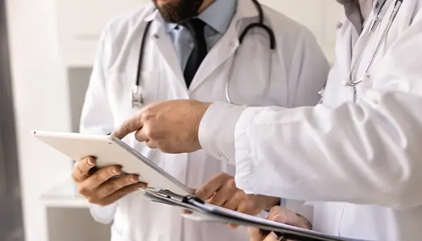Two doctors in white coats review a tablet and clipboard together in a clinical setting.