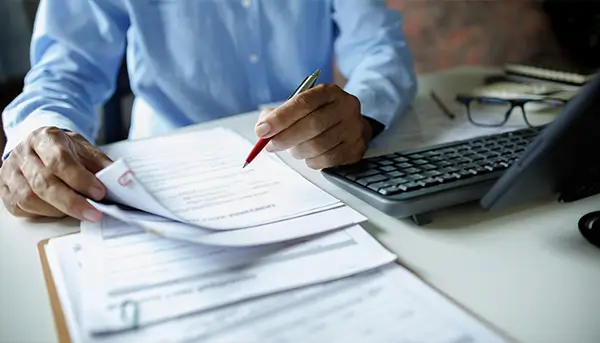 Person in a blue shirt signs documents at a desk with a red pen, laptop and glasses nearby.